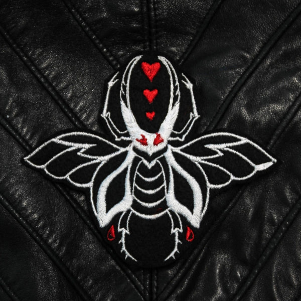 An embroidered patch of a large white beetle with wings on a black background. It has red eyes and there are three small red hearts in between its pincers. Shown on a leather jacket