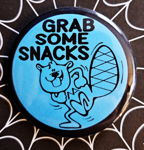 1.25” round button of an illustration of a cartoon beaver with the message “GRAB SOME SNACKS” in black on a neon blue background with black border 