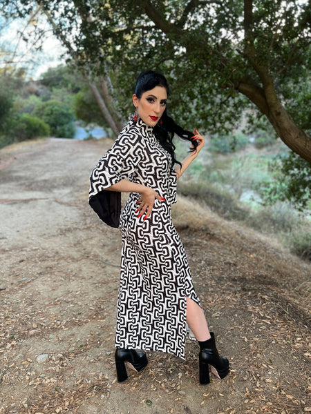 Model wearing maxi length dress in black and white concentric wavy psychedelic pattern. It has a mock neckline, fitted bodice, and long bell sleeves. Seen from side to display below the knee slit