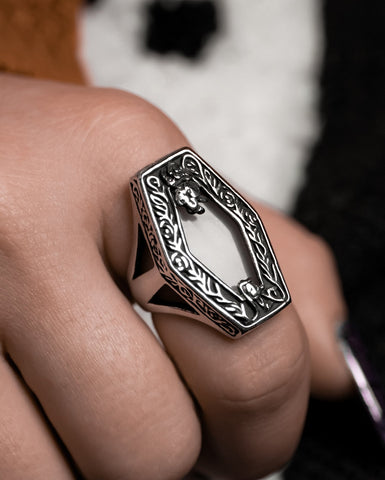 A model wearing a large ring with a hexagonal top decorated with filigree details and a large white semi translucent middle. Seen from the left hand side 