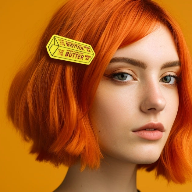 Laser-cut acrylic hair clip in the shape of a yellow stick of butter with black and red painted label details. Shown worn by a model