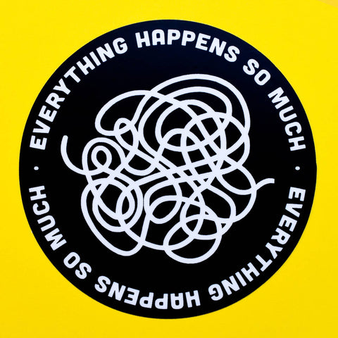 Round sticker with with a black background and the words “Everything happens so much” repeating around the border of the sticker. There is an abstract swirled shape in white in the middle