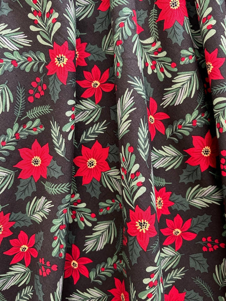 Mid length cotton dress with surplice neckline, cap sleeves, wide cummerbund style waistband, and full skirt. In a red and green holly, poinsettia, and mistletoe pattern on a black background. Shown in close up of pattern detail
