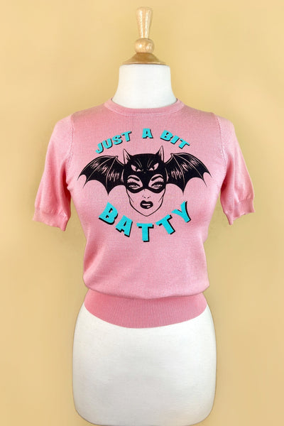 pink crew neck short sleeved sweater with retro woman wearing a bat mask and “Just a Bit Batty” written in bright blue. Shown on dress form