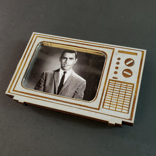 A refrigerator magnet in the shape of a vintage television in matte gray with brown details laser-cut Draftboard layered over black acrylic base, featuring Rod Sterling, the host of the classic tv show The Twilight Zone, on the screen in black and white
