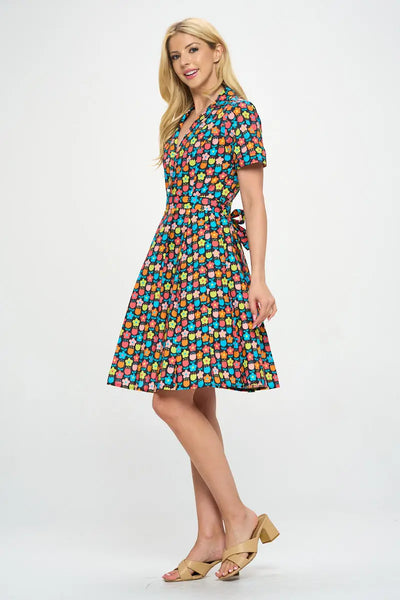 Model wearing floral shirtwaist dress in a stylized 60s/70s pattern of daisies and tulips in bright yellow, green, blue, and pink on a black background. It has short sleeves and a princess seamed bodice and notched v-neck collar. The skirt is pleated and knee length. Shown from 3/4 angle to display self sash belt