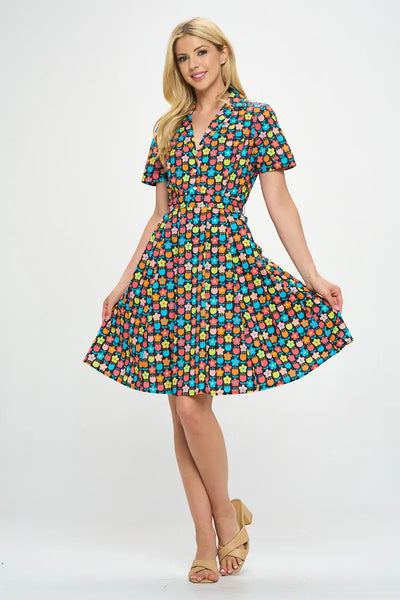 Model wearing floral shirtwaist dress in a stylized 60s/70s pattern of daisies and tulips in bright yellow, green, blue, and pink on a black background. It has short sleeves and a princess seamed bodice and notched v-neck collar. The skirt is pleated and knee length. Shown from front with skirt held up