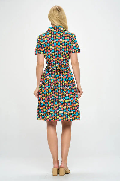 Model wearing floral shirtwaist dress in a stylized 60s/70s pattern of daisies and tulips in bright yellow, green, blue, and pink on a black background. It has short sleeves and a princess seamed bodice and notched v-neck collar. The skirt is pleated and knee length. Shown from back