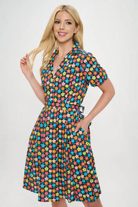 Model wearing floral shirtwaist dress in a stylized 60s/70s pattern of daisies and tulips in bright yellow, green, blue, and pink on a black background. It has short sleeves and a princess seamed bodice and notched v-neck collar. The skirt is pleated and knee length. The model has one hand in a pocket and is shown from the front