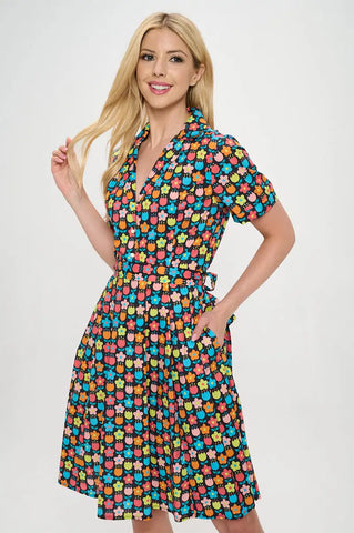 Model wearing floral shirtwaist dress in a stylized 60s/70s pattern of daisies and tulips in bright yellow, green, blue, and pink on a black background. It has short sleeves and a princess seamed bodice and notched v-neck collar. The skirt is pleated and knee length. The model has one hand in a pocket and is shown from the front