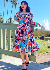 A model wearing a halter dress and bolero set in a bright blue, orange, pink, red, white, and black abstract tropical leaf pattern. It has a sweetheart neckline with exaggerated cuff details, cinched bodice, below the knee skirt, and adjustable halter tie neck. The bolero has short sleeves and a high mandarin style collar. Shown from the front
