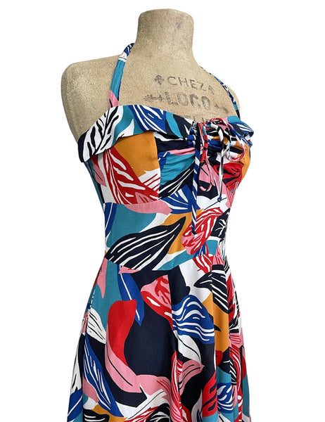 A halter dress in a bright blue, orange, pink, red, white, and black abstract tropical leaf pattern. It has a sweetheart neckline with exaggerated cuff details, cinched bodice, below the knee skirt, and adjustable halter tie neck. Shown on a dress form from a three quarter angle in close up