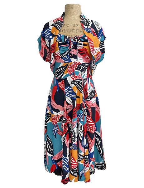 a halter dress and bolero set in a bright blue, orange, pink, red, white, and black abstract tropical leaf pattern. It has a sweetheart neckline with exaggerated cuff details, cinched bodice, below the knee skirt, and adjustable halter tie neck. The bolero has short sleeves and a high mandarin style collar. Shown from the front on a dress form 