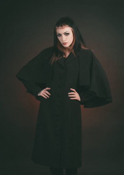 Model wearing black full length faux wool-blend coat with shawl collar and attached elbow-length cape. Shown with from the front