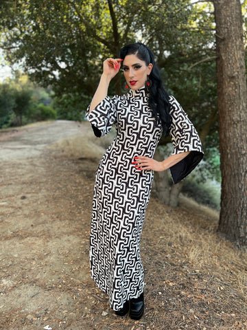 Model wearing maxi length dress in black and white concentric wavy psychedelic pattern. It has a mock neckline, fitted bodice, and long bell sleeves. Seen from fromt