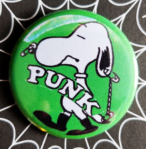 1.25” round pinback button of Snoopy with safety pins in his nose and caption “PUNK” on a green background 