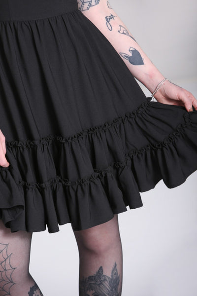 black mini dress featuring princess seamed fitted bodice with a lace edged angled v-neckline and velvet bow detail at the bust, sheer puffed short sleeves with elasticized cuffs, and a gathered full above the knee length skirt finished with a double row of flounces. showing close up detail of skirt on a model.