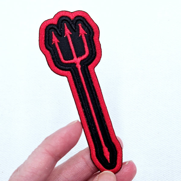 Black and red embroidered patch of a red devil’s pitchfork on a black background with red border. Shown held for scale