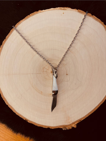 An 18” silver metal chain link necklace with a mini pocket knife charm in the shape of a white marbled enameled leg wearing a silver high heel shoe. Shown flat on a tree stump