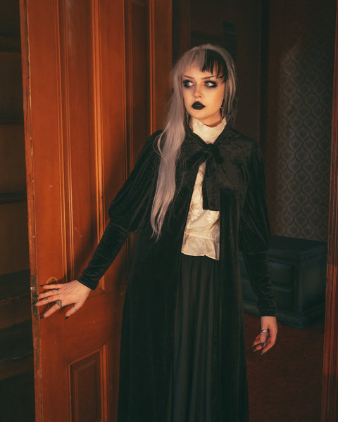 Model wearing black velvet duster with bow tied detail at the collar, slightly gathered shoulders, and Juliet sleeves. Shown from the front in close up
