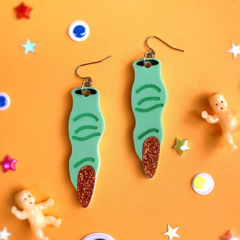 A pair of bright green laser-cut acrylic dangle earrings in the shape of a pair of witch’s fingers. With contrasting green detailing and glittery red fingernails. On an orange background 