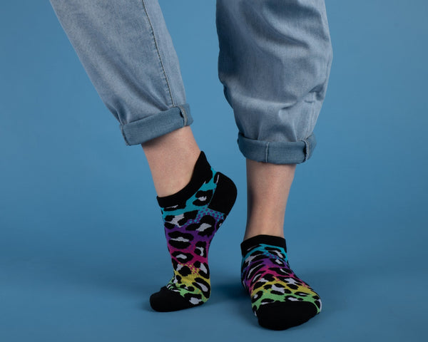 Neon blue, purple, pink, yellow, and green gradient leopard print ankle socks with black cuffs, toes, and heels. Shown worn by a model on a blue background 