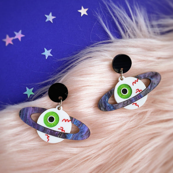 Laser-cut acrylic drop earrings in the shape of a pair of bloodshot green irised eyeballs being surrounded by swirled glittery purple and black rings, in the style of Saturn. Connects to ears by round black charm. Shown lying flat on a blue star covered background with white faux fur
