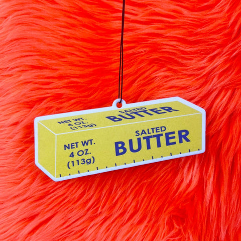 Butter shaped heavy cardstock vanilla scented air freshener with elastic hanging loop. Shown on a red faux fur background 