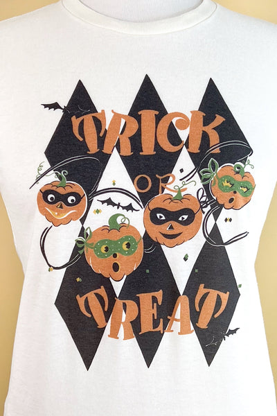 ivory short sleeved t shirt with an illustration of 4 jack-o’-lanterns wearing masks surrounded by bats in front of a harlequin diamond pattern with the message “TRICK OR TREAT” written in orange capital letters. Seen on a dress form in closeup