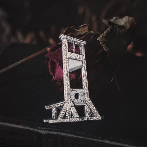 Antiqued silver metal enamel pin of a guillotine on a black background in front of a dead rose