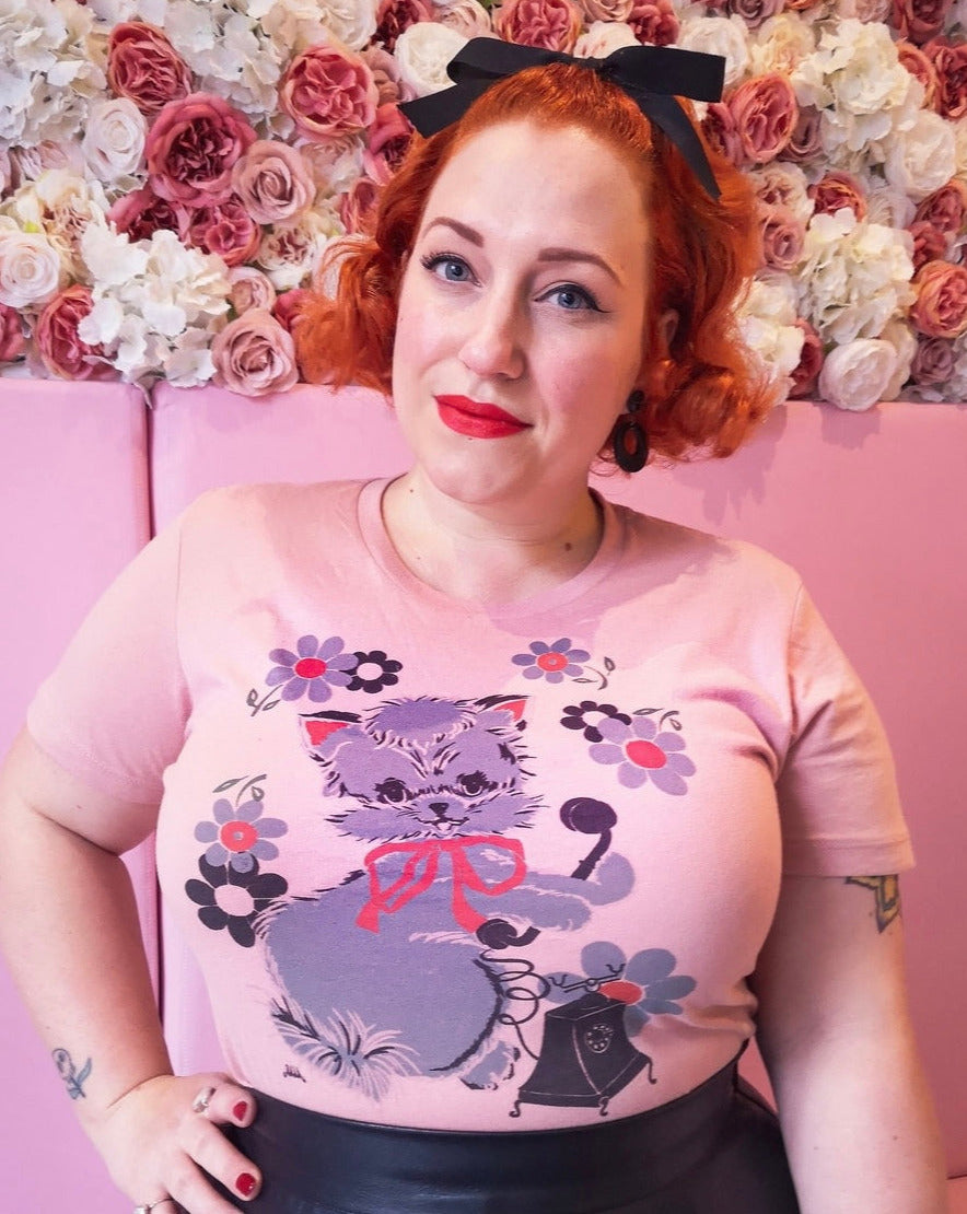 A model wearing a pink fitted t shirt with a print of a grey kitten wearing a pink bow holding the receiver of a rotary phone and surrounded by matching pink amends grey flowers