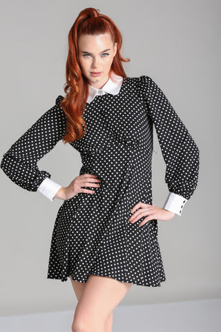 Model wearing black and white polka dot mini dress with bright white Peter Pan collar and matching white cuffs. Dress has long full sleeves with slightly puffed shoulders and a star-shaped silver metal button with pearl embellishments at the center of the collar. Skirt is flared and above the knee. Seen from the front
