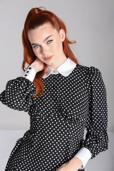 Model wearing black and white polka dot mini dress with bright white Peter Pan collar and matching white cuffs. Dress has long full sleeves with slightly puffed shoulders and a star-shaped silver metal button with pearl embellishments at the center of the collar. Skirt is flared and above the knee. Seen from the front in close up