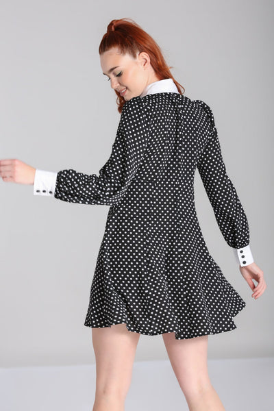 Model wearing black and white polka dot mini dress with bright white Peter Pan collar and matching white cuffs. Dress has long full sleeves with slightly puffed shoulders and a star-shaped silver metal button with pearl embellishments at the center of the collar. Skirt is flared and above the knee. Seen from the back