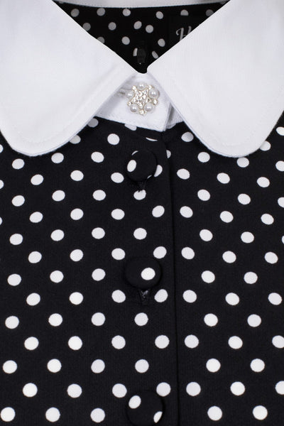 black and white polka dot mini dress with bright white Peter Pan collar and matching white cuffs. Dress has long full sleeves with slightly puffed shoulders and a star-shaped silver metal button with pearl embellishments at the center of the collar. Skirt is flared and above the knee. Seen in close up of collar and button detail at bust