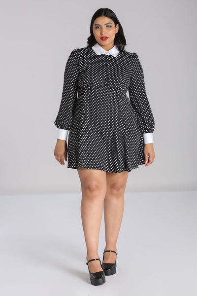Plus size model wearing black and white polka dot mini dress with bright white Peter Pan collar and matching white cuffs. Dress has long full sleeves with slightly puffed shoulders and a star-shaped silver metal button with pearl embellishments at the center of the collar. Skirt is flared and above the knee. Seen from the front 