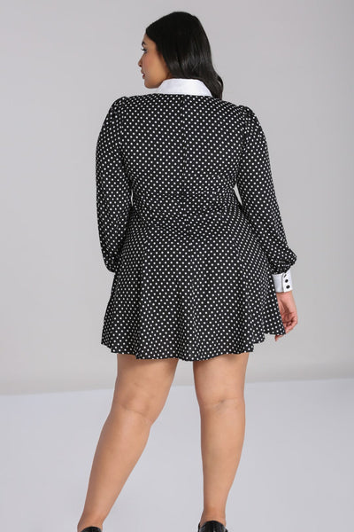 Plus size model wearing black and white polka dot mini dress with bright white Peter Pan collar and matching white cuffs. Dress has long full sleeves with slightly puffed shoulders and a star-shaped silver metal button with pearl embellishments at the center of the collar. Skirt is flared and above the knee. Seen from the back