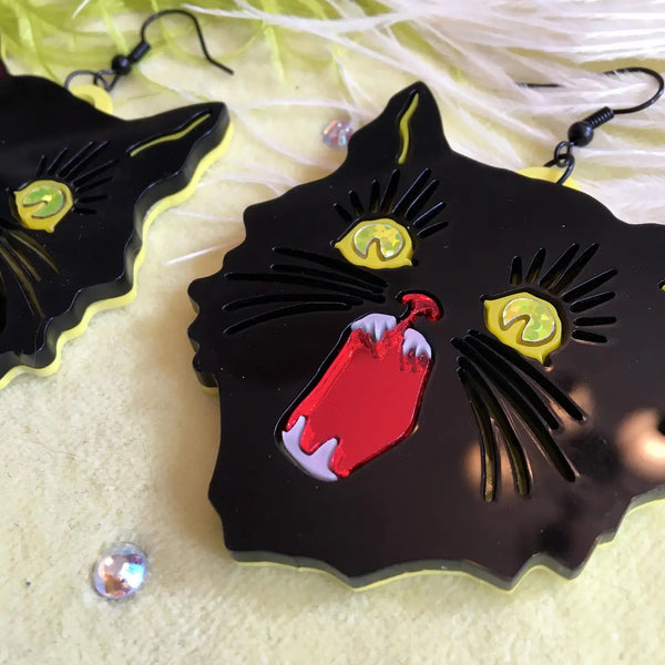 Dangle earrings in the shape of pair of black cats reminiscent of vintage Halloween decorations crafted out of layered laser-cut acrylic: black, mirrored red mouth, pearly iridescent teeth and yellow glitter foil eyes with a solid neon yellow back. Seen in close up