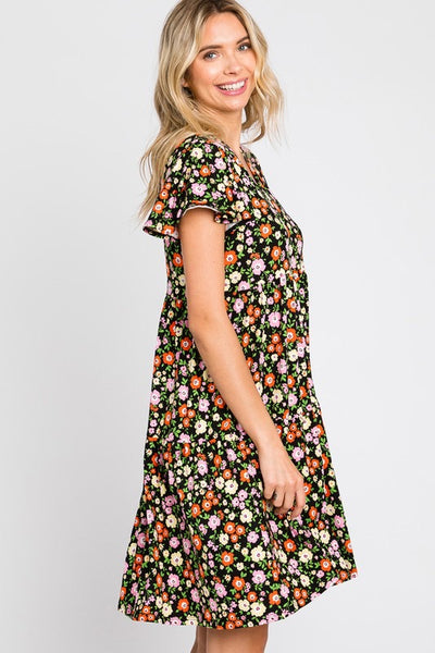 Knit dress in a red, pink, yellow, and green floral pattern on a black background. The dress has a shallow v-neck and short flutter sleeves with a gathered at the waist tiered skirt that ends just above the knee. Shown on a model from the side