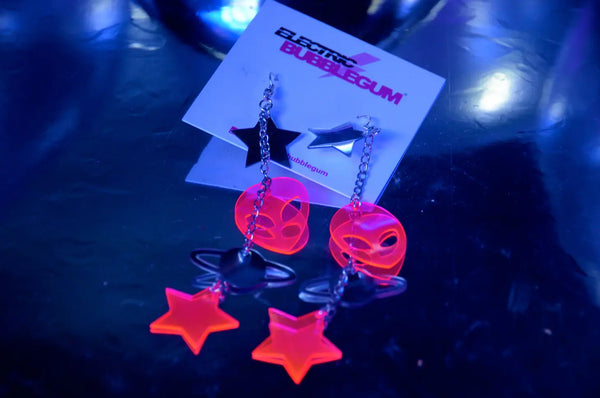 A pair of dangle earrings made of pieces of neon pink and silver chrome vinyl in alien, star, and planet shapes connected by pieces of silver metal chain. Shown on their card packaging under a UV light to display the glow