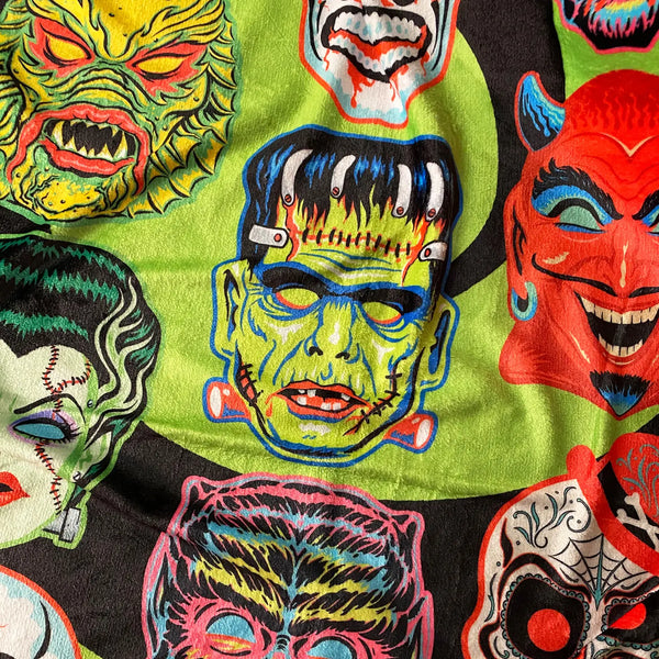 Throw blanket with all-over vibrantly colored print of retro Halloween monster masks on a swirled black and bright green background. Shown in close up to display fabric texture 