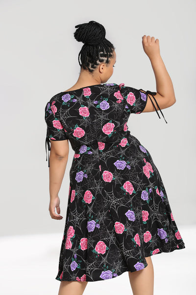Plus-size model wearing a mid length rayon dress in a stylized white spiderweb with purple and pink rose pattern on a black background. It has keyhole and tie details at both the slight v-neckline and the short sleeves. The bodice is slightly gathered under the bust and the skirt is flared just below the knee. Shown from the back 