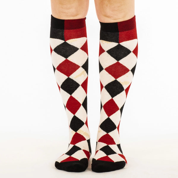 knee socks in a black, red, and cream colored harlequin pattern- with faux-antiqued yellowed age spots throughout each cream colored diamond in the pattern. Shown on a model from the front