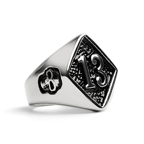 Shiny stainless steel signet style ring depicting the number 13 inside of a diamond shape on the top of the ring with a small skull on either side. Shown from the side