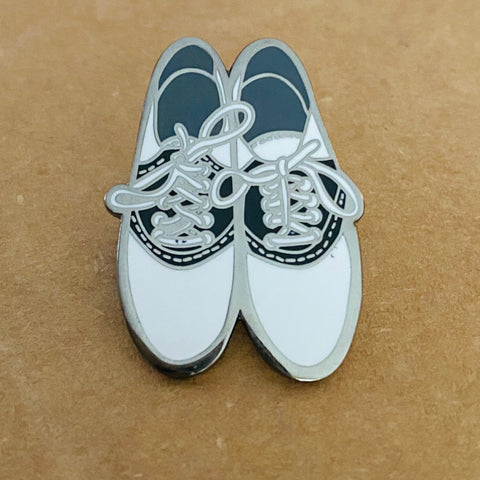 A silver enamel pin in the shape of a pair of black and white saddle shoes with white laces tied in bows