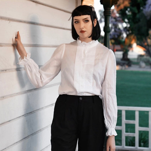 Model wearing a white button down shirt with high ruffled collar and cuffs. It has pin-tuck detail down the front of the shirt and down the sides of both puffed full sleeves.