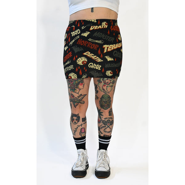 A black stretch knit mini skirt with an all-over pattern of bats, skeleton hands, dripping red blood, and eyeballs with the words “DEATH”, “GHOUL”, “MONSTER”, “HORROR” written in vintage movie poster style fonts in red, grey, and beige yellow. Seen on a model from the waist down from the front