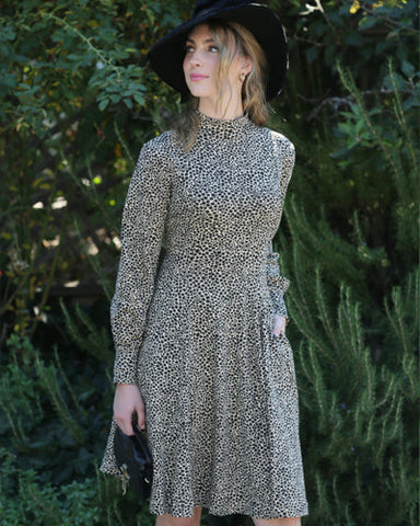 Model wearing a cotton spandex jersey dress with long sleeves and a mock turtleneck collar in a black ditsy floral pattern on a tan background. It has slightly puffed bishop style sleeves and a skirt that ends just below the knee