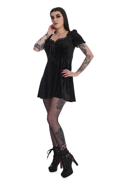 Model wearing a black stretch velvet mini dress with an all over embossed damask pattern of chandeliers, roses, candles, and crescent moons. It has lace trim at the bust and shoulders of the square neckline and a lace up notched detail at the bust. The skirt hits above the knee. Shown from the side