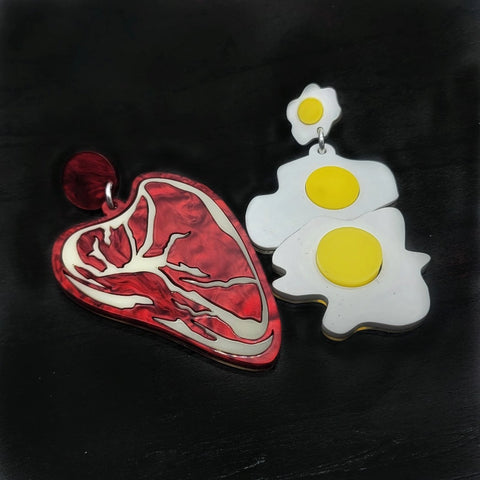 A pair of laser cut acrylic drop earrings in the shape of a rare ribeye steak with marbled red acrylic and two sunny side up eggs. Shown flat on a black background 